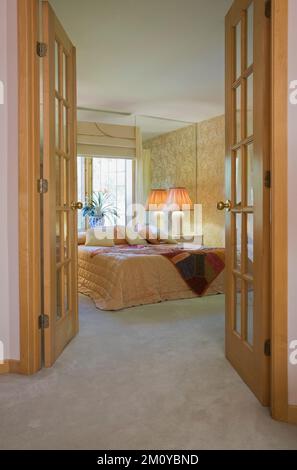 Kingsize-Bett im Hauptschlafzimmer mit großem Wandspiegel durch geöffnete glastüren in gemischt dekoriertem und architektonischem Stil Haus. Stockfoto