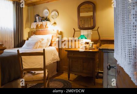 Antiker Schaukelstuhl neben Einzelbett im Gästezimmer im 1977 gebauten Nachbau des alten 1800er Jahre Canadiana Cottage Stil Blockhaus. Stockfoto