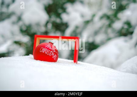 Fröhliche Weihnachten im Hintergrund. Rote Weihnachtskugel in einem Rahmen im Schnee in einem Winter verschneiten Wald. Weihnachtstapete. Winterferien Stockfoto