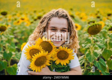 Porträt einer jungen Frau mit lockigem Haar, die den Sommer im Sonnenblumenfeld bei Sonnenuntergang genießt. Wunderschöner Sonnenuntergang. Lächle Emotionen. Stockfoto