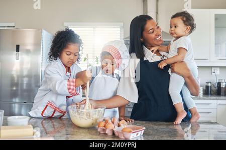 Wir machen Pfannkuchen, um den neuen Zahn zu feiern. Eine Mutter, die ihr Baby hält, während sie mit ihren beiden Kindern backt. Stockfoto