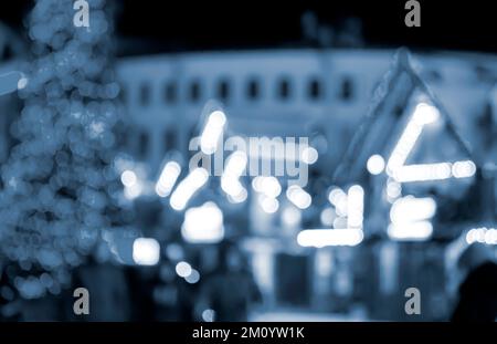 Unscharfer Hintergrund. Im Winter laufen die Leute auf dem Marktplatz. Silhouetten von Menschen, die in der Nähe von Häusern spazieren, dekoriert mit leuchtender Beleuchtung. Weiße Bokeh-Spots von leuchtenden Hauslichtern Blaue Farbe Stockfoto