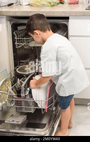 Kleiner Junge, der die Spülmaschine auflädt und Verantwortung in der Küche übernimmt. Geschirrspüler sparen Zeit und Wasser. Stockfoto