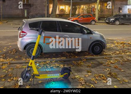 Eriwan, Armenien - 3. Dezember 2022: Ein elektrischer Roller neben einem geparkten Auto in der Abendstraße. Gelb gefallenes trockenes Laub auf der Straße. Stockfoto