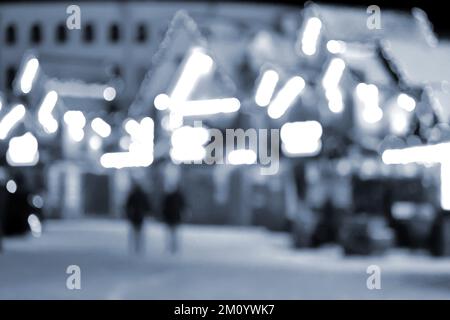 Unscharfer Hintergrund. In der Winternacht laufen die Leute in der Stadt herum. Schwarze Silhouetten von Menschen, die in der Nähe von Häusern spazieren, dekorierte, leuchtende Beleuchtung. Weiße Bokeh-Spots von leuchtenden Hauslichtern Blaue Farbe Stockfoto