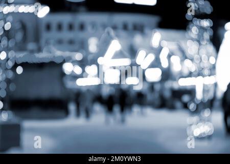 Unscharfer Hintergrund. In der Winternacht laufen die Leute in der Stadt herum. Schwarze Silhouetten von Menschen, die in der Nähe von Häusern spazieren, dekorierte, leuchtende Beleuchtung. Weiße Bokeh-Spots von leuchtenden Hauslichtern Blaue Farbe Stockfoto