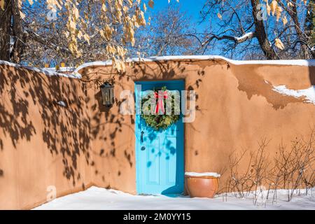 Verschneite Winterszene mit Weihnachtskranz an der türkisfarbenen Tür in der lehmwand in Santa Fe, New Mexico Stockfoto