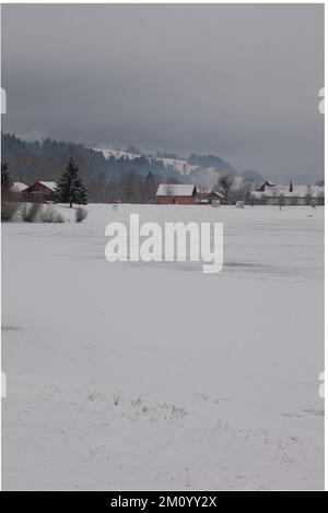 Der Himmel ist bedeckt, die Hügel sind weiß mit Schnee, der See ist gefroren und teilweise mit Schnee bedeckt, die Häuser haben Dächer weiß mit Schnee. Stockfoto