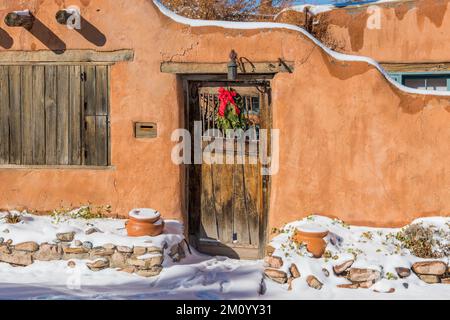 Verschneite Winterszene mit Weihnachtskranz auf einer rustikalen Holztür in der lehmwand in Santa Fe, New Mexico Stockfoto