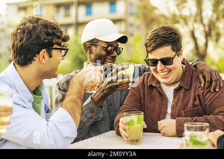 Vielfältige Gruppe von 20s jungen Freunden, die im Pub Cafe im Freien scherzten und lachten - multiethnische junge Leute, die im Tter sitzen und Mojito-Cocktails trinken Stockfoto
