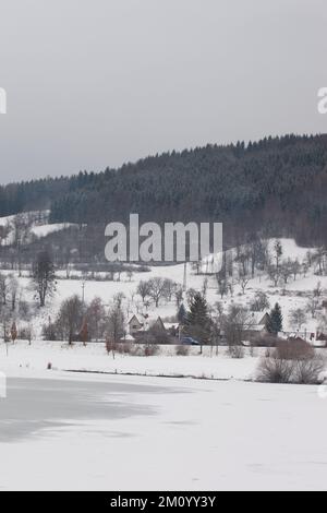 Der Himmel ist bedeckt, die Hügel sind weiß mit Schnee, der See ist gefroren und teilweise mit Schnee bedeckt, die Häuser haben Dächer weiß mit Schnee. Stockfoto