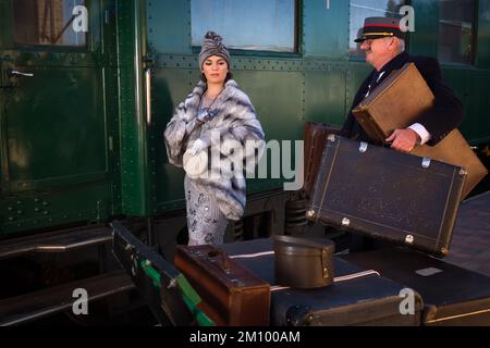 Nachstellungsszene auf dem Bahnsteig in der Nähe eines authentischen 1927 First Class Zugwaggons, wo eine schicke 1920er Dame auf ihr Gepäck wartet Stockfoto