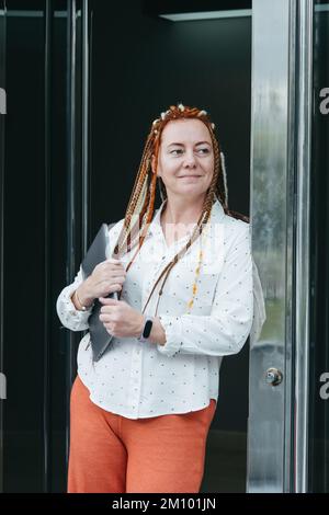 Eine Frau mit Dreadlocks und einem Laptop, die sich gegen die Haustür lehnt und auf ihre Uhr schaut Stockfoto