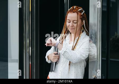 Eine Frau mit Dreadlocks und einem Laptop, die sich gegen die Haustür lehnt und auf ihre Uhr schaut Stockfoto