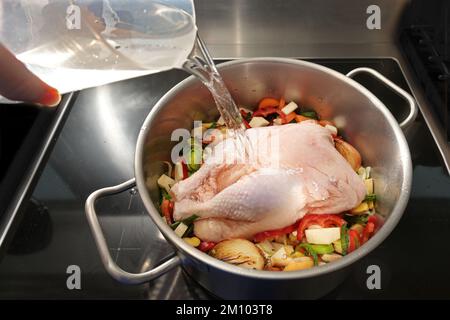 Ganzes rohes Hähnchen in einem Topf mit gehacktem Gemüse wird mit kaltem Wasser gegossen, um eine gesunde Suppe gegen Erkältung und Grippe zu kochen, ausgewählter Fokus, schmaler Tag Stockfoto