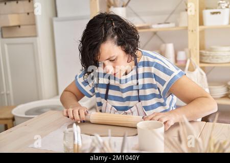 Frau in blau gestreiftem Hemd knete Ton in einer Töpferwerkstatt. Frau lernt, Lehm zu arbeiten. Handwerksklasse. Stockfoto