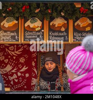 Eine junge Verkäuferin steht hinter dem Tresen mit einem Geschäftsmann im Weihnachtsmarkt-Kiosk und verkauft Backkartoffeln Stockfoto