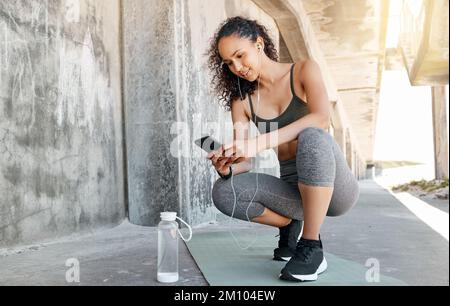 Wie sieht meine Gesundheitsstatistik aus? Aufnahme einer attraktiven jungen Frau in der ganzen Länge, die sich während ihres Workouts im Freien in der Hocke ruht und ihr Handy benutzt. Stockfoto