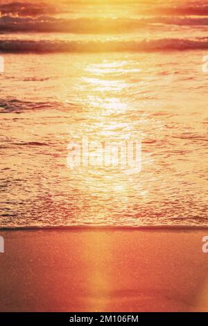 Sonnenuntergang über dem Meer. Riplpe Meer Ozean Wasseroberfläche mit kleinen Wellen. Ozean Wasser Schaum Spritzwasser waschen Sandstrand. Vertikales Foto Stockfoto