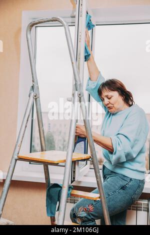 Glücklich Schüchtern Reifen Frau Reinigung Fenster. Kaukasische Frau Im Mittleren Alter. Ältere Frau Reinigung Apartment. Konzept Für Aktive Senioren. Hausfrau. Konzept Von Stockfoto