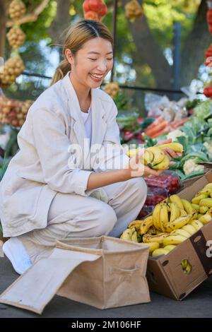 Bananen, Einkaufen und asiatische Frau auf einem Markt im Freien, die köstliche und gesunde Früchte kauft. Mehlbananen, Produkte und Frauen aus Japan, die Obst bei kaufen Stockfoto