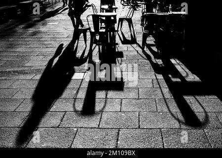 Lange Schatten von Stühlen, Tischen und Figuren in Schwarz und Weiß. Dramatische Hintergrundbeleuchtung mit Schatten, die direkt auf die Kamera zugehen. Schattenkonzept Stockfoto