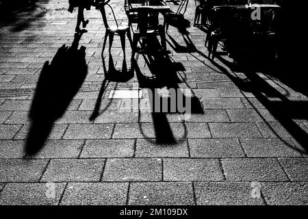 Lange Schatten von Stühlen, Tischen und Figuren in Schwarz und Weiß. Dramatische Hintergrundbeleuchtung mit Schatten, die direkt auf die Kamera zugehen. Schattenkonzept Stockfoto