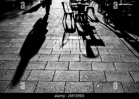 Lange Schatten von Stühlen, Tischen und Figuren in Schwarz und Weiß. Dramatische Hintergrundbeleuchtung mit Schatten, die direkt auf die Kamera zugehen. Schattenkonzept Stockfoto