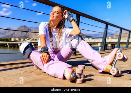 Foto einer fröhlichen jungen glücklichen Frau Tragen Sie an sonnigen Tagen weißes T-Shirt und eine Hose mit Glockenboden und rosafarbenen, pastellfarbenen Rollschuhen im Freien Stockfoto