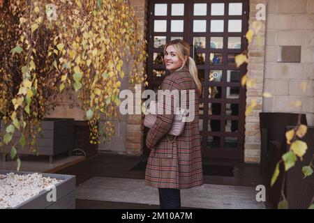 Porträt einer stilvollen Frau mit einem schönen Lächeln, langen blonden Haaren, modischem Herbstlook, brauner karierter Jacke. Das junge Model ist dabei Stockfoto