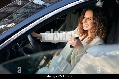 Attraktive junge Frau, die eine Tasse Kaffee hält und lächelt, während sie ein Elektrofahrzeug fährt. Fröhliche Geschäftsfrau in eleganter weißer Jacke, die Kaffee im Auto trinkt. Stockfoto