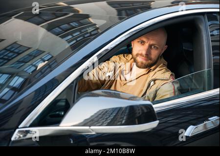 Porträt eines erwachsenen Mannes, der ein schwarzes Auto fährt. Ein Mann mit Bart sitzt im Auto und legt seine Hände auf das Lenkrad mit abgesenktem Fenster in der Seitentür. Stockfoto