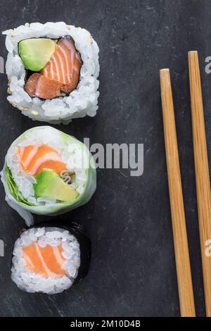 Von oben sehen Sie drei verschiedene Lachs Makis auf schwarzem Steinhintergrund mit Stäbchen daneben. Kopierraum. Makroaufnahme. Stockfoto