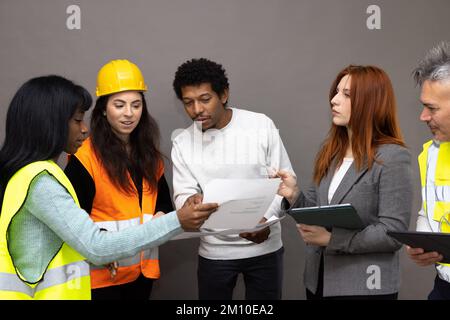 Gemischte Gruppe von Baustellenarbeitern, die das Projekt beobachten und diskutieren. Männer und Frauen verschiedener ethnischer Herkunft, die Warnwesten tragen Stockfoto