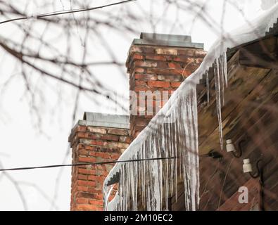 Gefährliche Eiszapfen hängen am Dachrand, im Winter oder Frühling. Holzwand eines alten Holzhauses mit Fenstern. Große Eiszapfen in glatten, schönen Reihen. Bewölkter Wintertag, weiches Licht. Stockfoto
