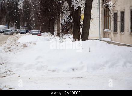 Ein kleiner Schneewetter vor dem Hintergrund einer Straße mit Bäumen. Auf der Straße liegt weißer Schnee in hohen Haufen. Urbane Winterlandschaft. Bewölkter Wintertag, weiches Licht. Stockfoto