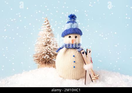 Weihnachtskomposition eines gestrickten Schneemanns mit blauem Hut und Schal mit Skiern auf dem Hintergrund eines kleinen Weihnachtsbaums Stockfoto
