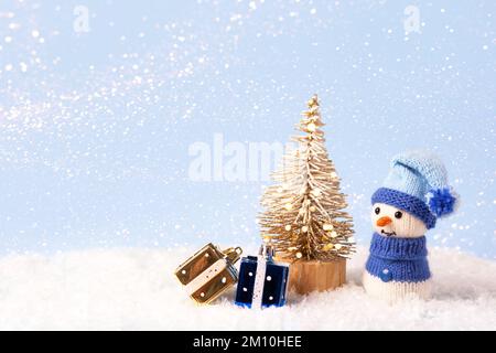 Weihnachtsmärchen-Komposition eines gestrickten Schneemanns mit blauem Hut und Pullover. Kleiner Weihnachtsbaum mit zwei gelben und blauen Geschenken Stockfoto