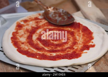 Fügen Sie Tomatensoße mit einem Löffel auf Pizzateig auf einer Schaufel auf einem Holzbrett hinzu, Nahaufnahme Stockfoto