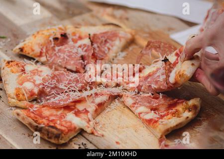 Hände nehmen Stücke oder Scheiben Pizza mit Tomatensoße, Mozzarella, Parmesankäse und Salami-Fleisch von einem Holzbrett Stockfoto