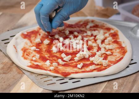 Hände mit Kunststoffhandschuhen Fügen Sie frischen Mozzarella auf eine Pizza mit Tomatensoße auf einem Holzbrett mit Zutaten hinzu, Nahaufnahme Stockfoto