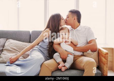 Mom und Dad küssen sich, während das Kind in die Kamera schaut. Glückliche Eltern, die zu Hause auf der Couch vor dem Fenster sitzen. Familienbeziehung und Liebe Stockfoto