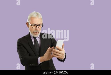 Schlechte Nachrichten. Ein überraschter Senior-Mann, der auf dem Smartphone-Bildschirm eine schockierende Nachricht liest. Posieren auf lila Studiohintergrund. Stockfoto