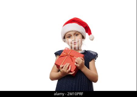 Hübsches lächelndes kleines Mädchen mit Weihnachtsmann-Hut, posieren mit niedlicher Geschenkbox mit Weihnachtsgeschenk auf weißem Hintergrund Stockfoto