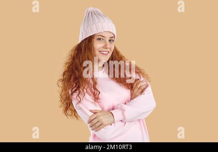 Lächelndes rothaariges Mädchen mit lockigem, langem Haar in pinkfarbenem Sweatshirt und Hut auf beigefarbenem Hintergrund. Stockfoto