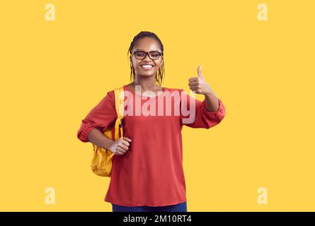 Glückliche afroamerikanische Schülerin mit Brille, die den Daumen nach oben zeigt, isoliert auf gelbem Hintergrund. Stockfoto