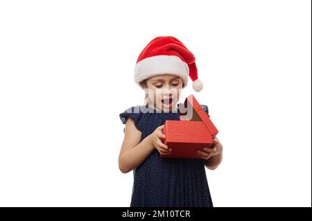 Ein überraschtes Kind mit Weihnachtsmannmütze, das Begeisterung ausdrückt, während es eine rote Geschenkbox mit niedlichem Weihnachtsgeschenk auspackt Stockfoto