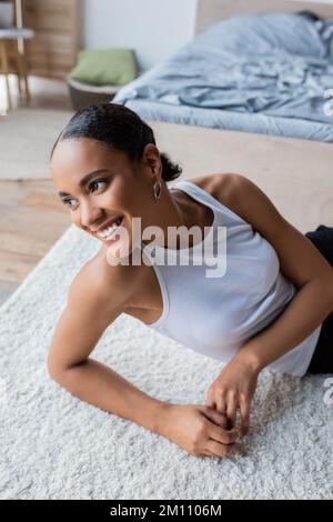 Junge und fröhliche afroamerikanische Frau im Tank, die auf dem Teppich sitzt, Stock Image Stockfoto