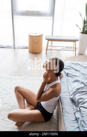 Blick von oben auf eine junge afroamerikanische Frau im Tank, die auf dem Teppich neben dem Bett sitzt, Stockbild Stockfoto