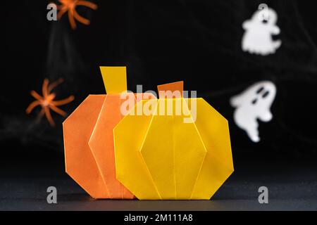 Origami Kürbisse aus Papier für die Halloween-Feiertage, vor dem Hintergrund fliegender Geister auf schwarzem Hintergrund, machen es selbst. Stockfoto